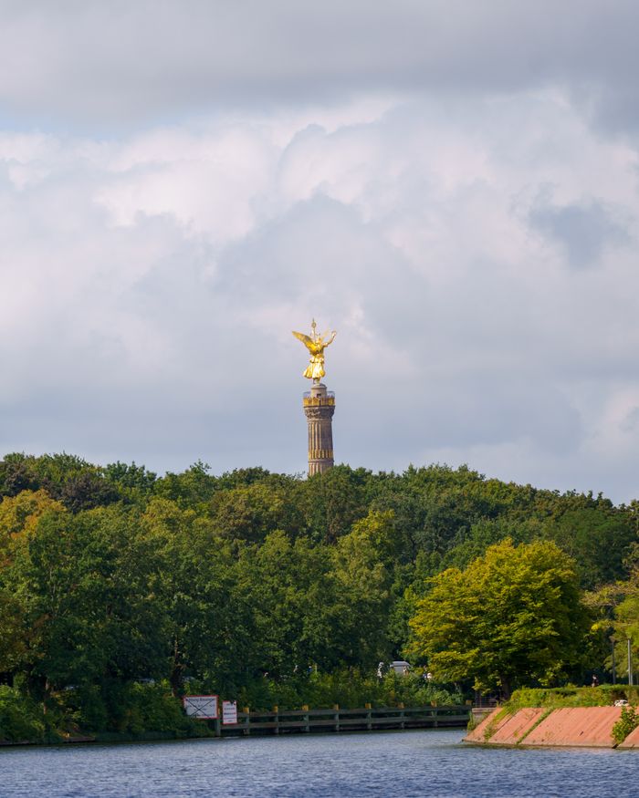 The iconic Berlin Victory Column peeks above lush green trees on a cloudy day.